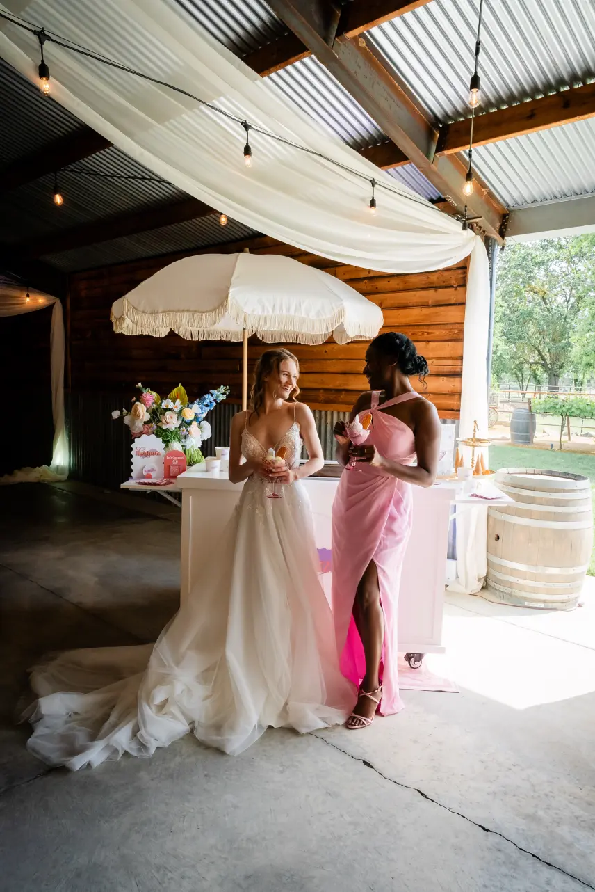 Two women in dresses holding gelato coupes beside the Blush Fizz Co. cart at a wedding celebration